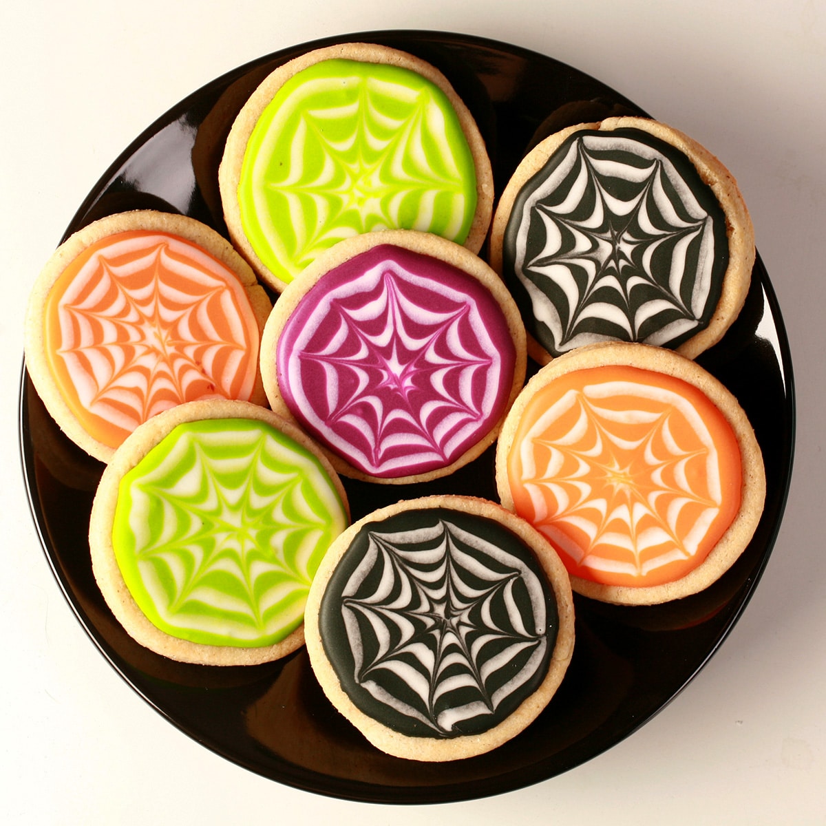 Close up view of a plate of round halloween cookies, frosted with orange, lime green, purple, and black individually. Each has a white stylized spider web design in the coloured background.