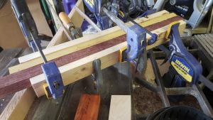 Several strips of different colored wood are held together with five clamps.
