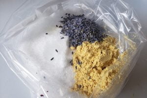 Close up view of a plastic baggie with 3 different substances in it - small purple lavender flowers, yellow mustard powder, and white epsom salt.