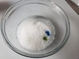 Close up photo of a glass bowl with salt in it. There are two spots of colour in the salt - a green spot and a blue spot.