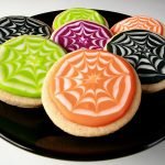 Close up view of a plate of round halloween cookies, frosted with orange, lime green, purple, and black individually. Each has a white stylized spider web design in the coloured background.