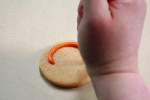 A hand in the foreground is applying orange frosting to a cookie