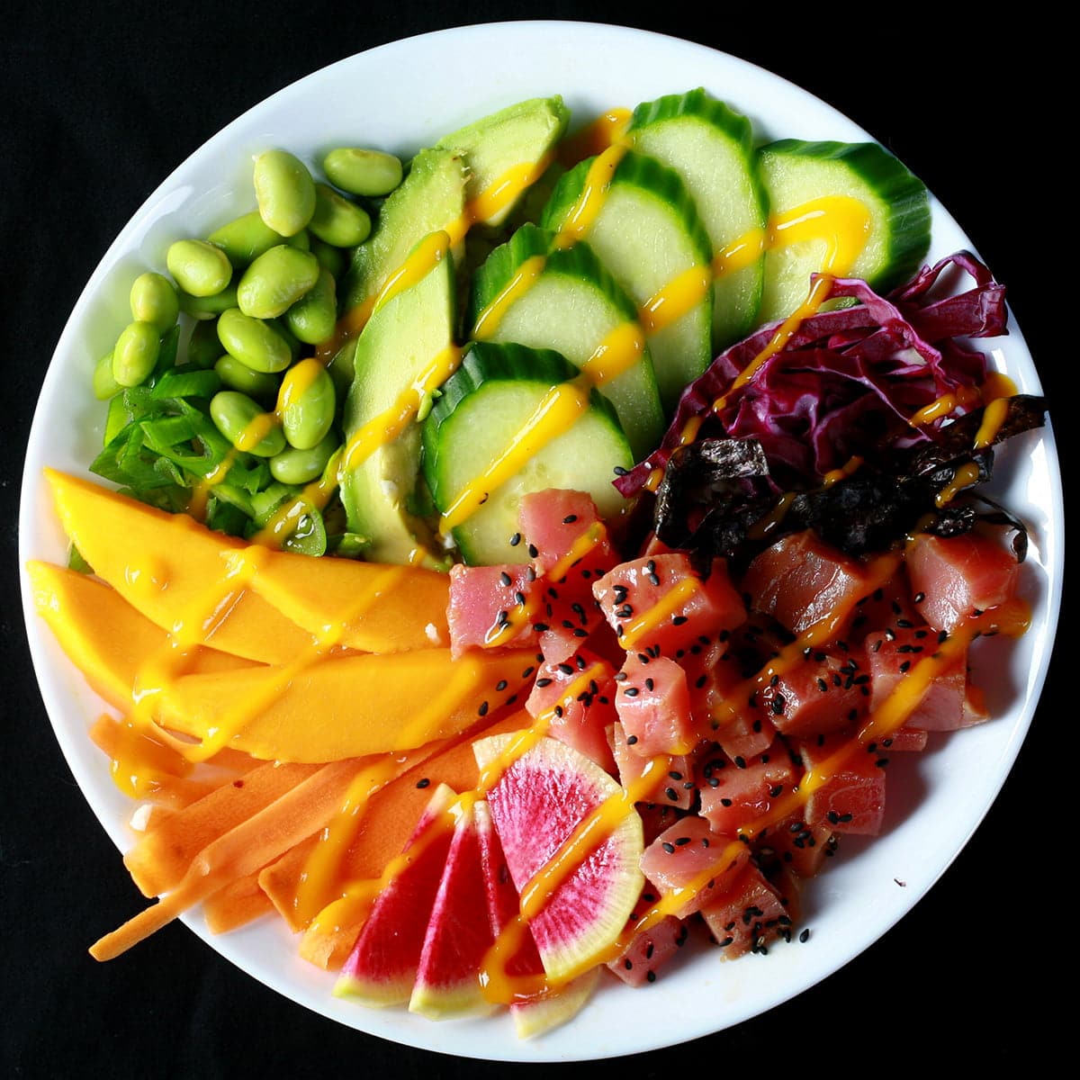 Close up view of a colourful bowl of tuna mango poke. There is a rainbow of vegetables and fruit, including purple cabbage, watermelon radish, carrots, mango, edamame, avocado, and cucumber