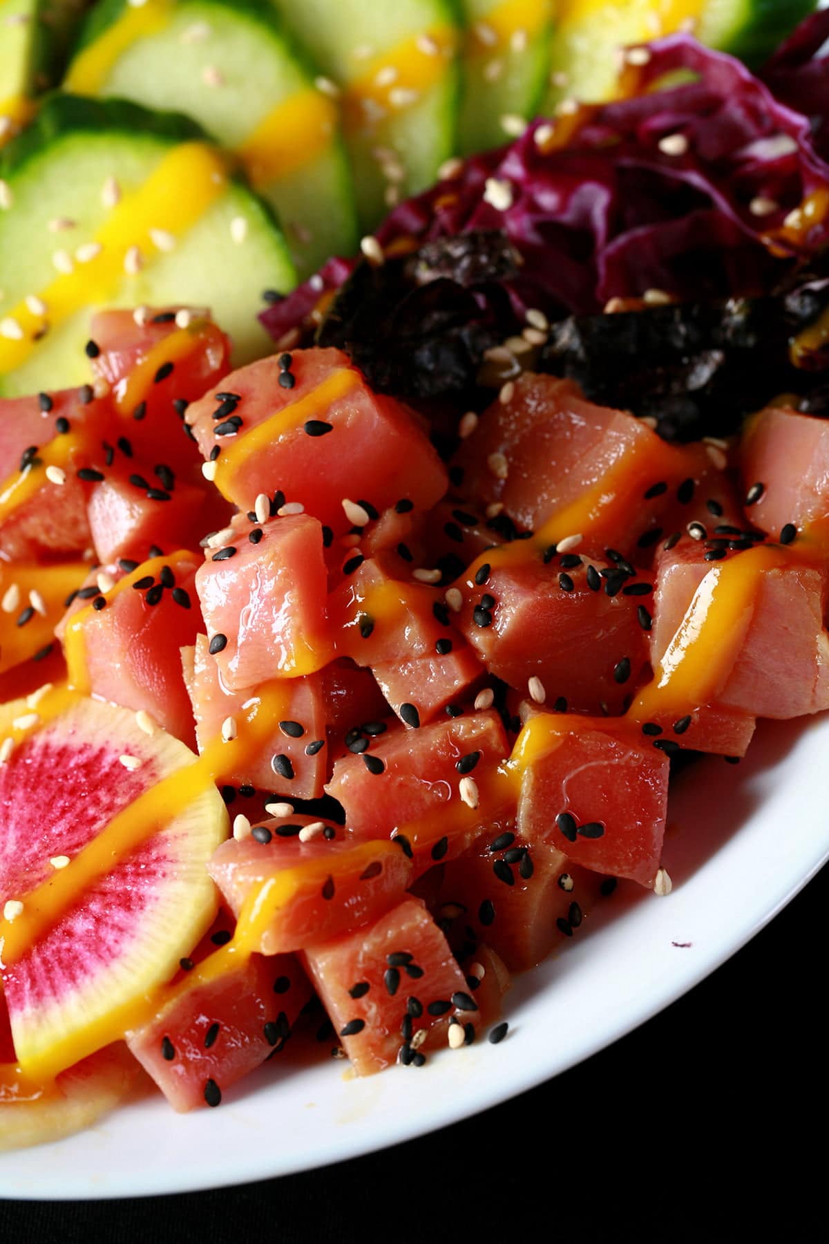 Close up view of a colourful bowl of tuna mango poke. There is a rainbow of vegetables and fruit, including purple cabbage, watermelon radish, carrots, mango, edamame, avocado, and cucumber
