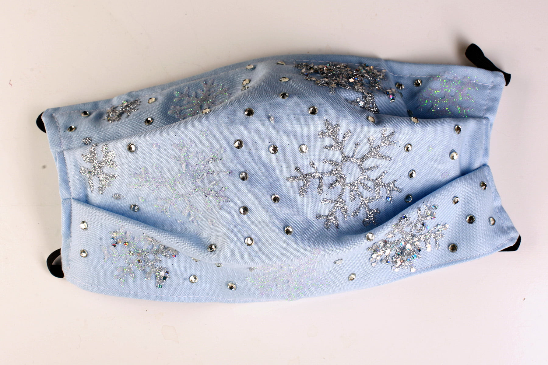 A light blue face mask with glittery white and silver snowflakes all over it.