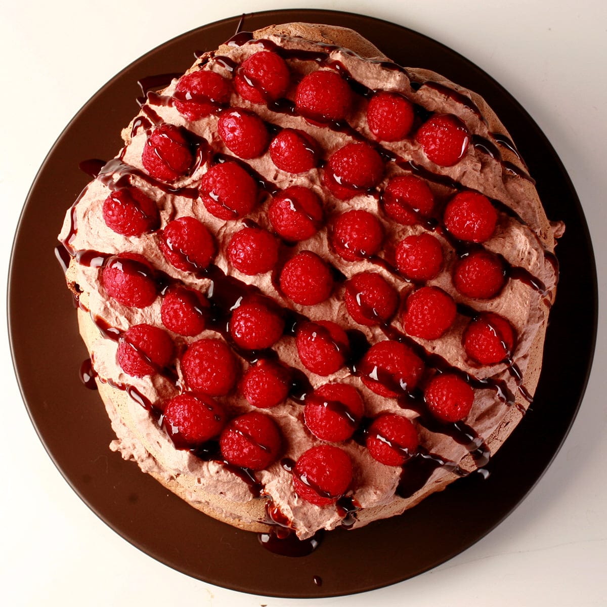 Top view of a chocolate raspberry pavlova. A round meringue is topped with chocolate whipped cream, raspberries, and chocolate sauce.