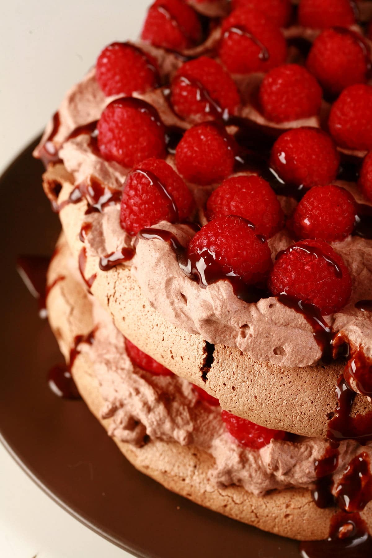 A double layered chocolate pavlova on a brown plate. Two layers of chocolate meringue are sandwiches with layers of chocolate whipped cream and raspberries, drizzled with chocolate sauce.