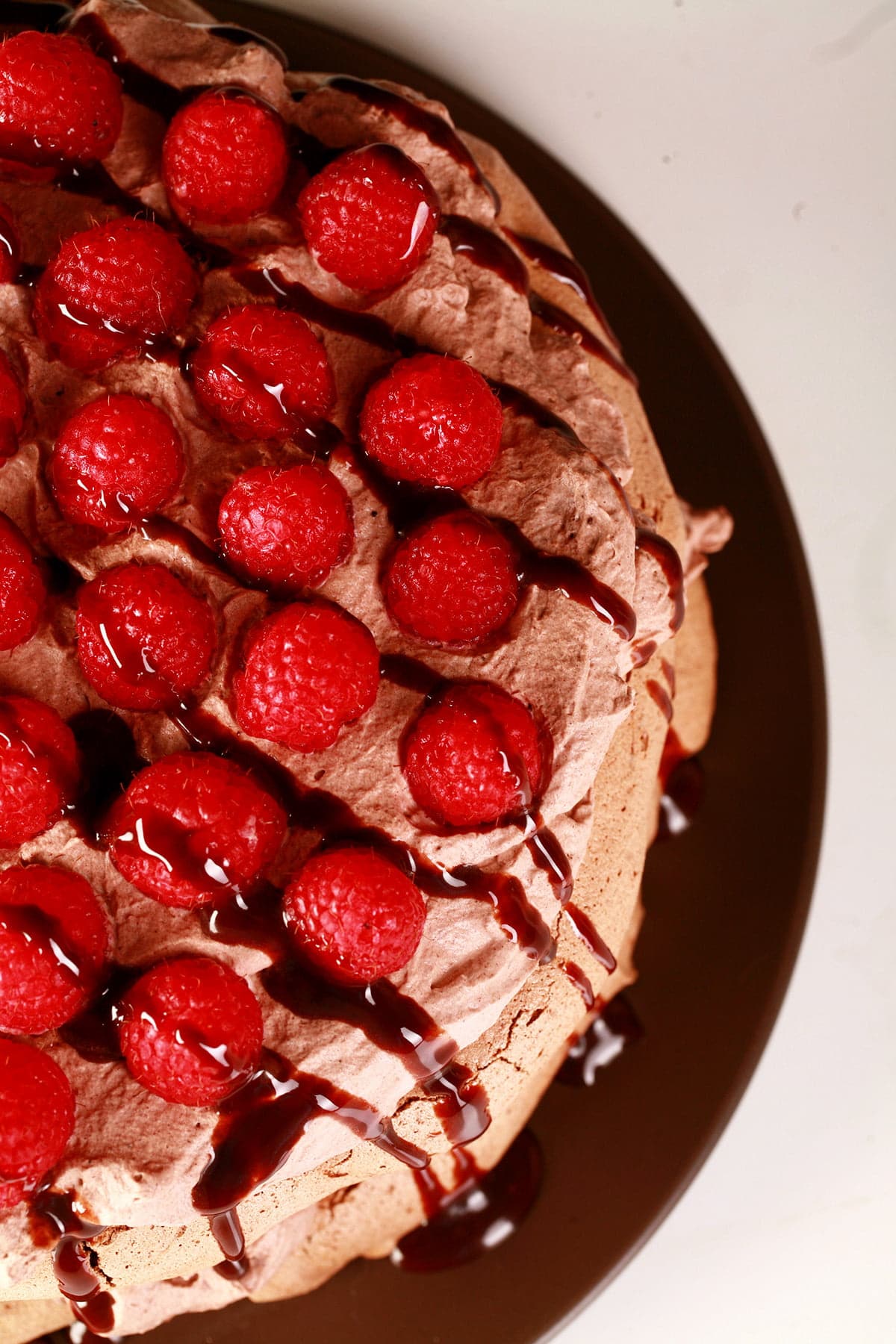 A double layered chocolate pavlova on a brown plate. Two layers of chocolate meringue are sandwiches with layers of chocolate whipped cream and raspberries, drizzled with chocolate sauce.