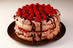 A double layered chocolate pavlova on a brown plate. Two layers of chocolate meringue are sandwiches with layers of chocolate whipped cream and raspberries, drizzled with chocolate sauce.