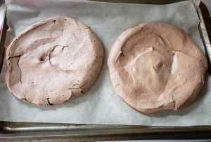 Two baked round chocolate meringues on a parchment lined baking sheet.