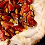 An ivory coloured Pavlova meringue round, piled with whipped cream, sliced figs, and pistachios. A drizzle of honey finishes it off.