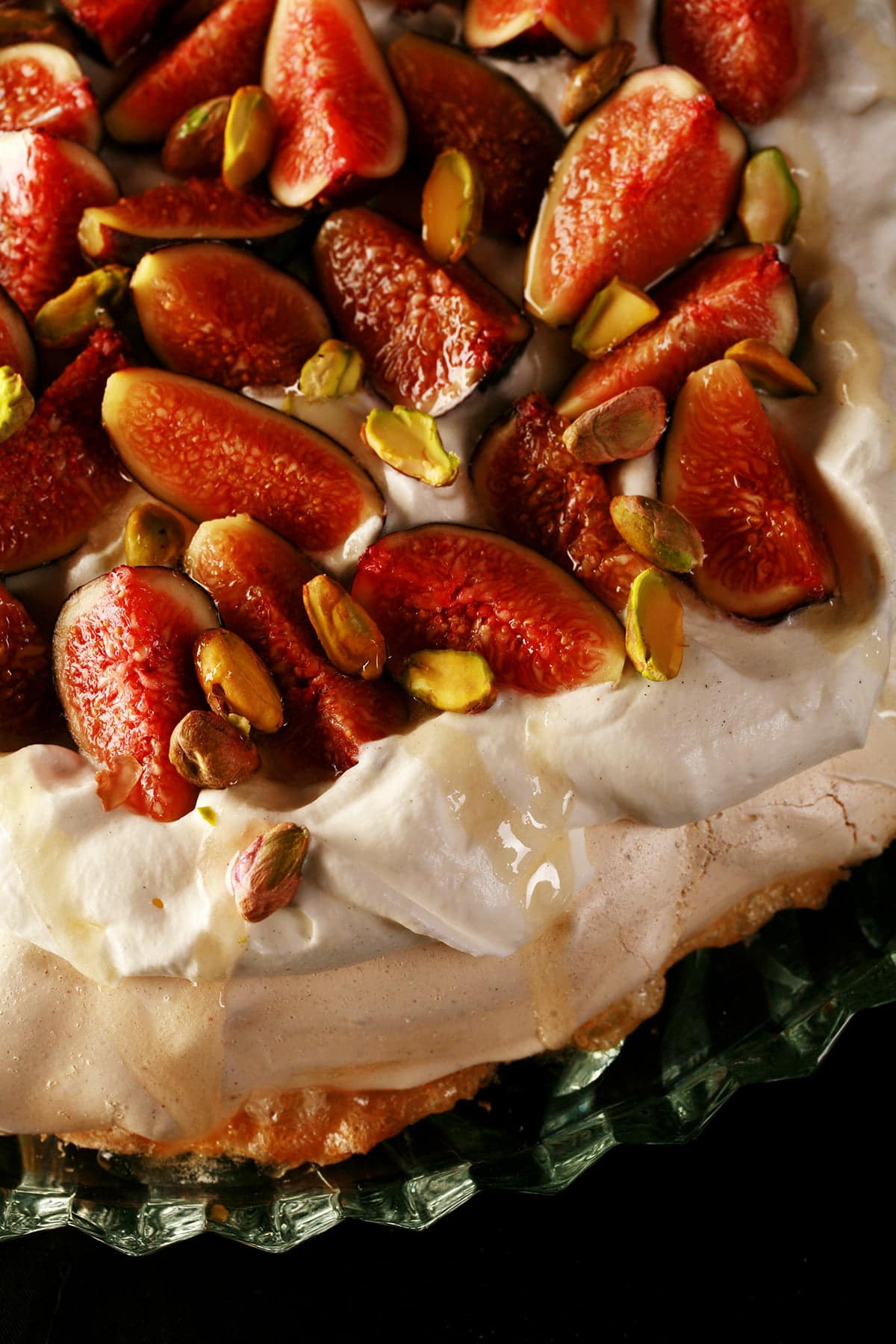 An ivory coloured Pavlova meringue round, piled with whipped cream, sliced figs, and pistachios. A drizzle of honey finishes it off.