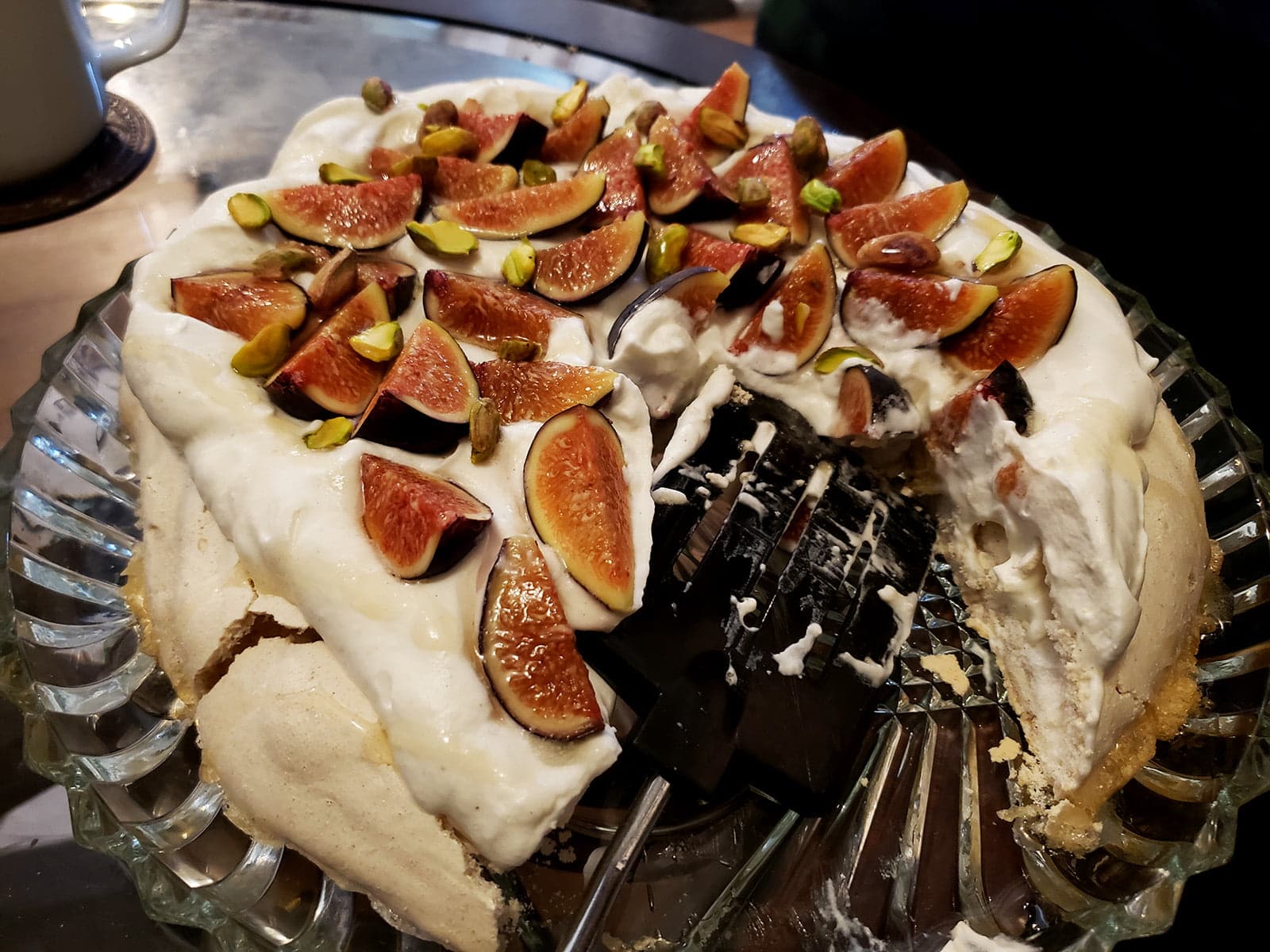 An ivory coloured Pavlova meringue round, piled with whipped cream, sliced figs, and pistachios. It's half eaten, on a glass serving plate.  A Cardamom fig pavlova.