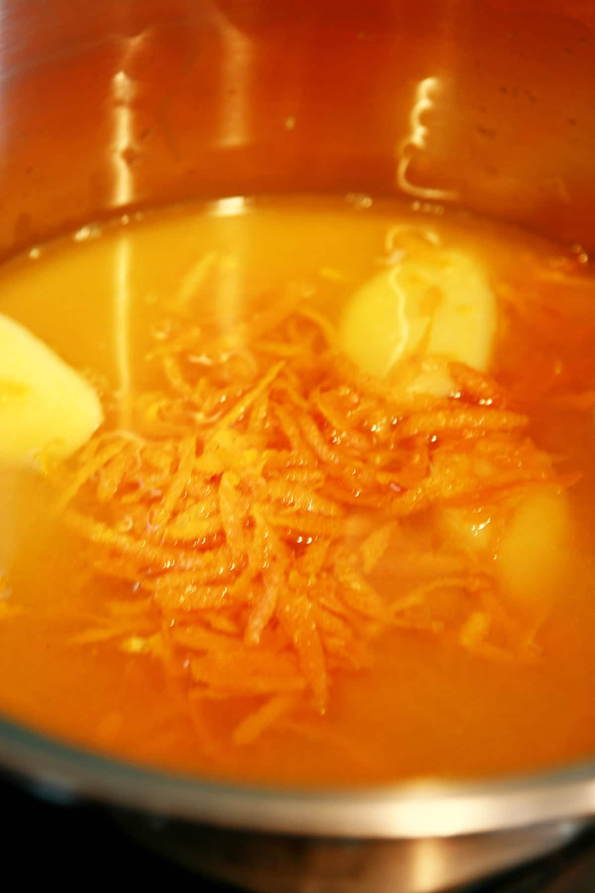 Close up view of the inside of a pot. It's got orange juice, orange zest, and slices of ginger in it.