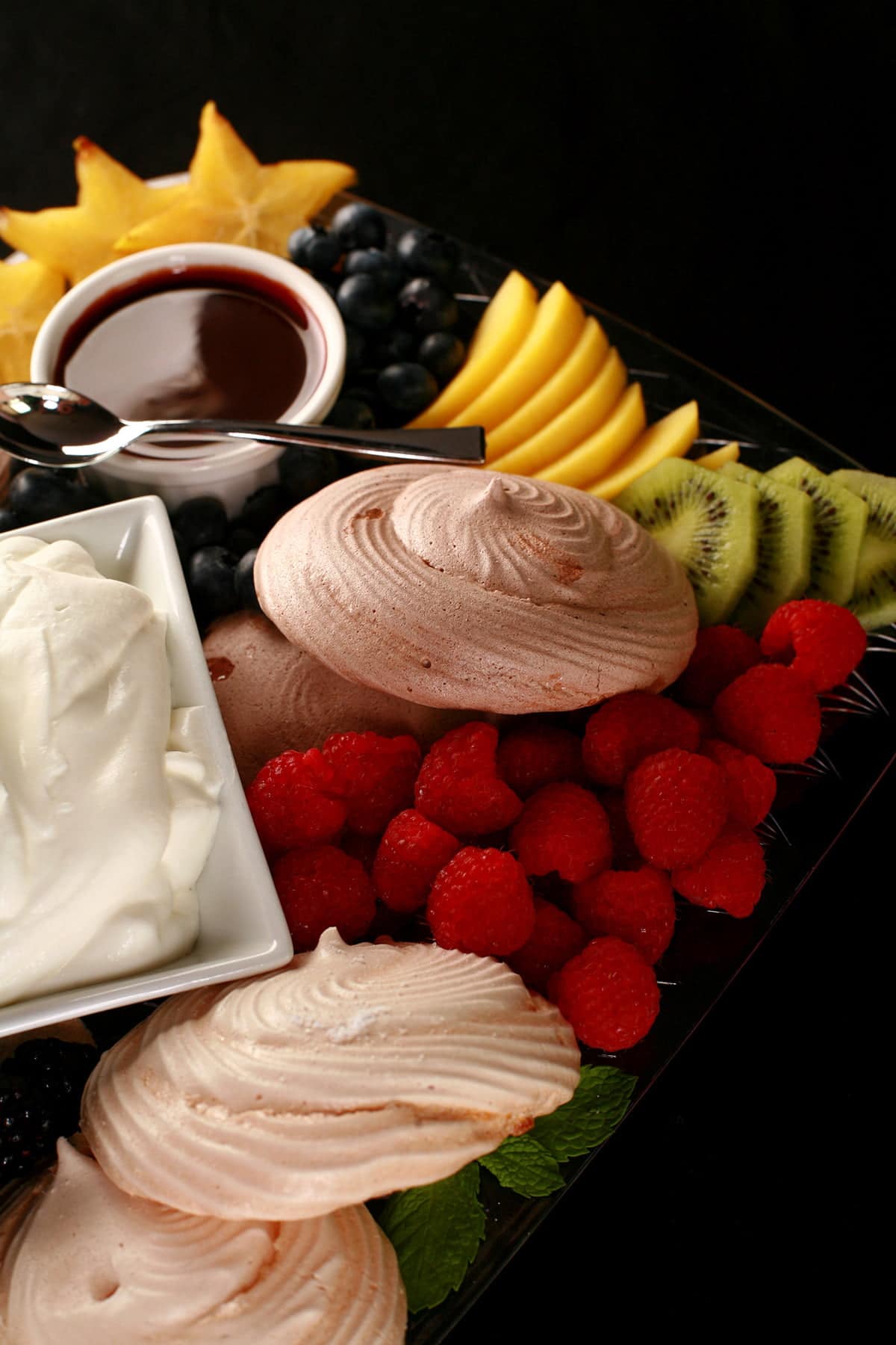 A Pavlova Grazing Board: A bowl of whipped cream and two little bowls of sauce - lemon curd and chocolate sauce - are surrounded by mini pavlova meringues and a selection of fresh fruits.