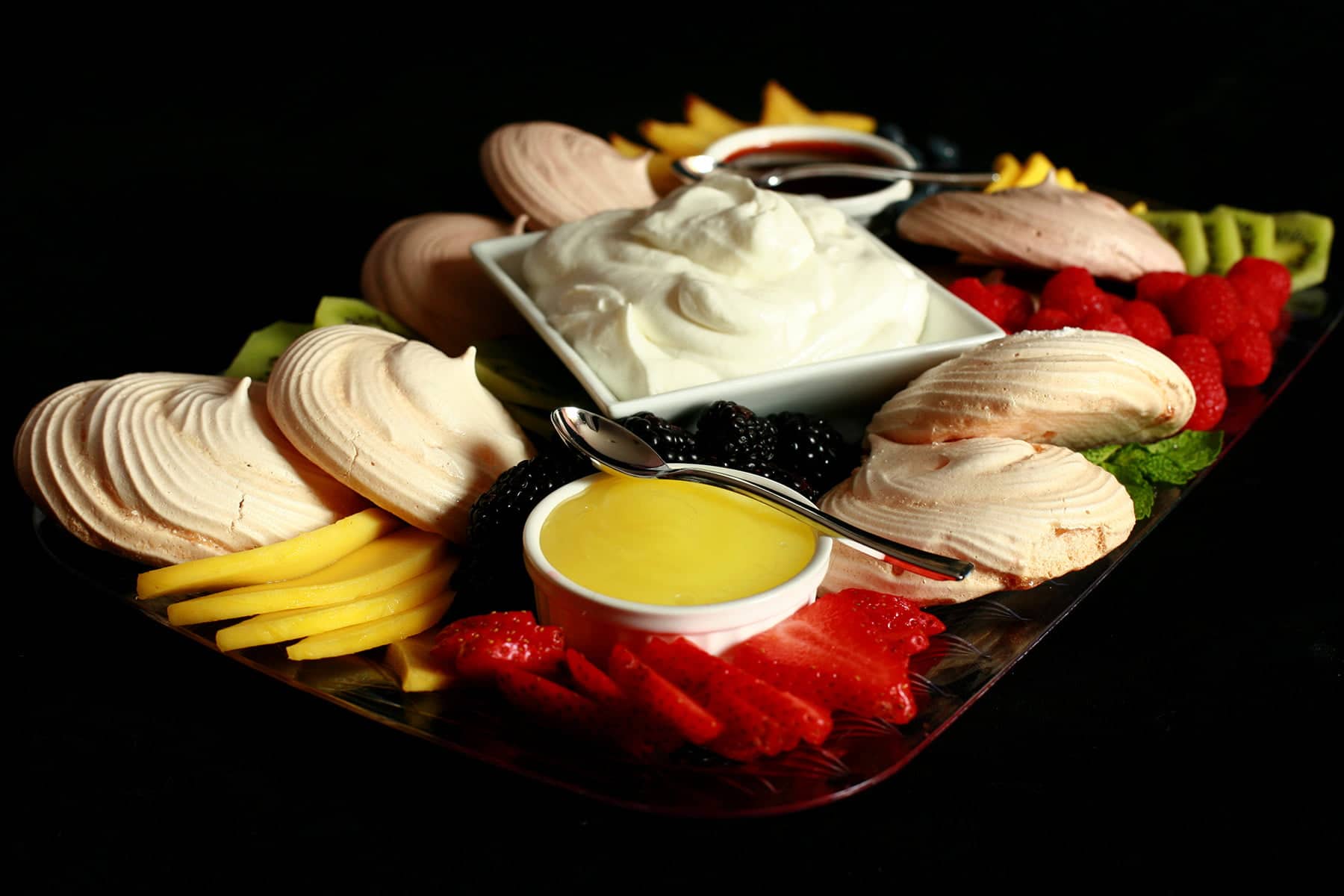 A Pavlova Dessert Charcuterie board: A bowl of whipped cream and two little bowls of sauce - lemon curd and chocolate sauce - are surrounded by mini pavlova meringues and a selection of fresh fruits.