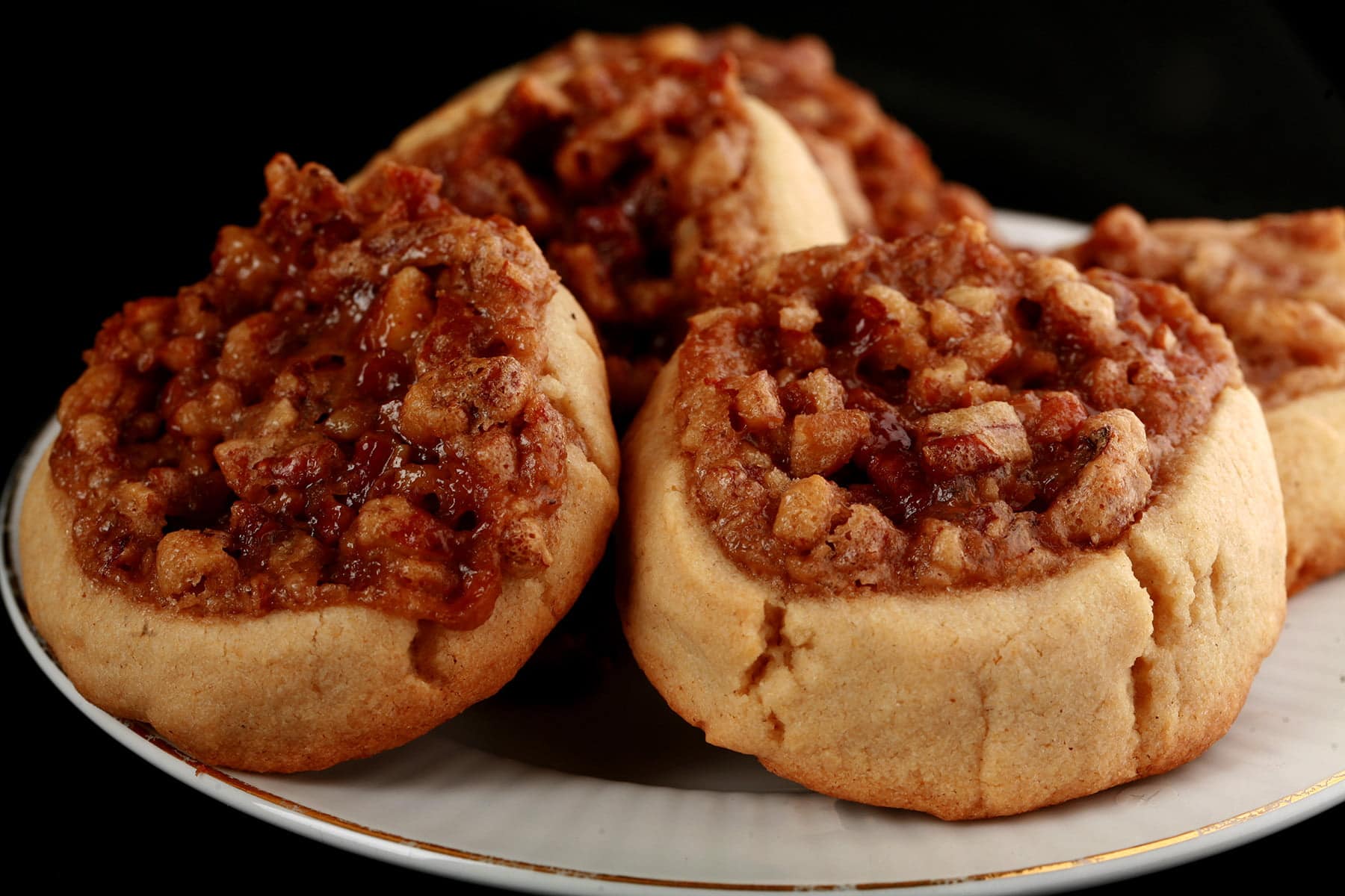A small plate with Pecan Pie Cookies on it. Each cookie is round and golden brown, with a center indentation filled with chopped pecans and caramelized brown sugar.