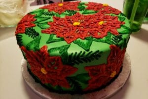 A large round cake covered in bright green fondant is covered in frosting piped to look like poinsettias. The design is mostly red and green, with accents of yellow.