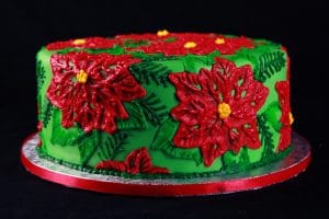 A large round cake covered in bright green fondant is covered in frosting piped to look like poinsettias. The design is mostly red and green, with accents of yellow.