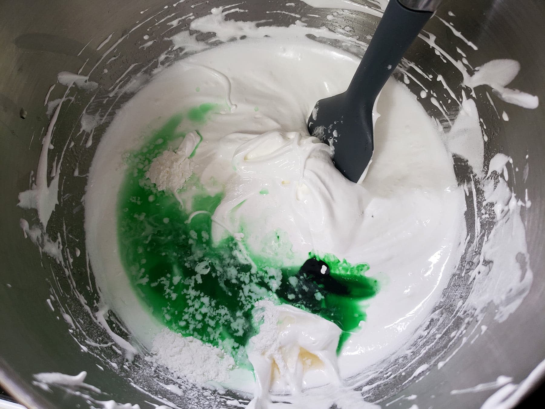 A bowl of stiff whipped meringue has corn starch, extract, vinegar, and food colouring added to it.