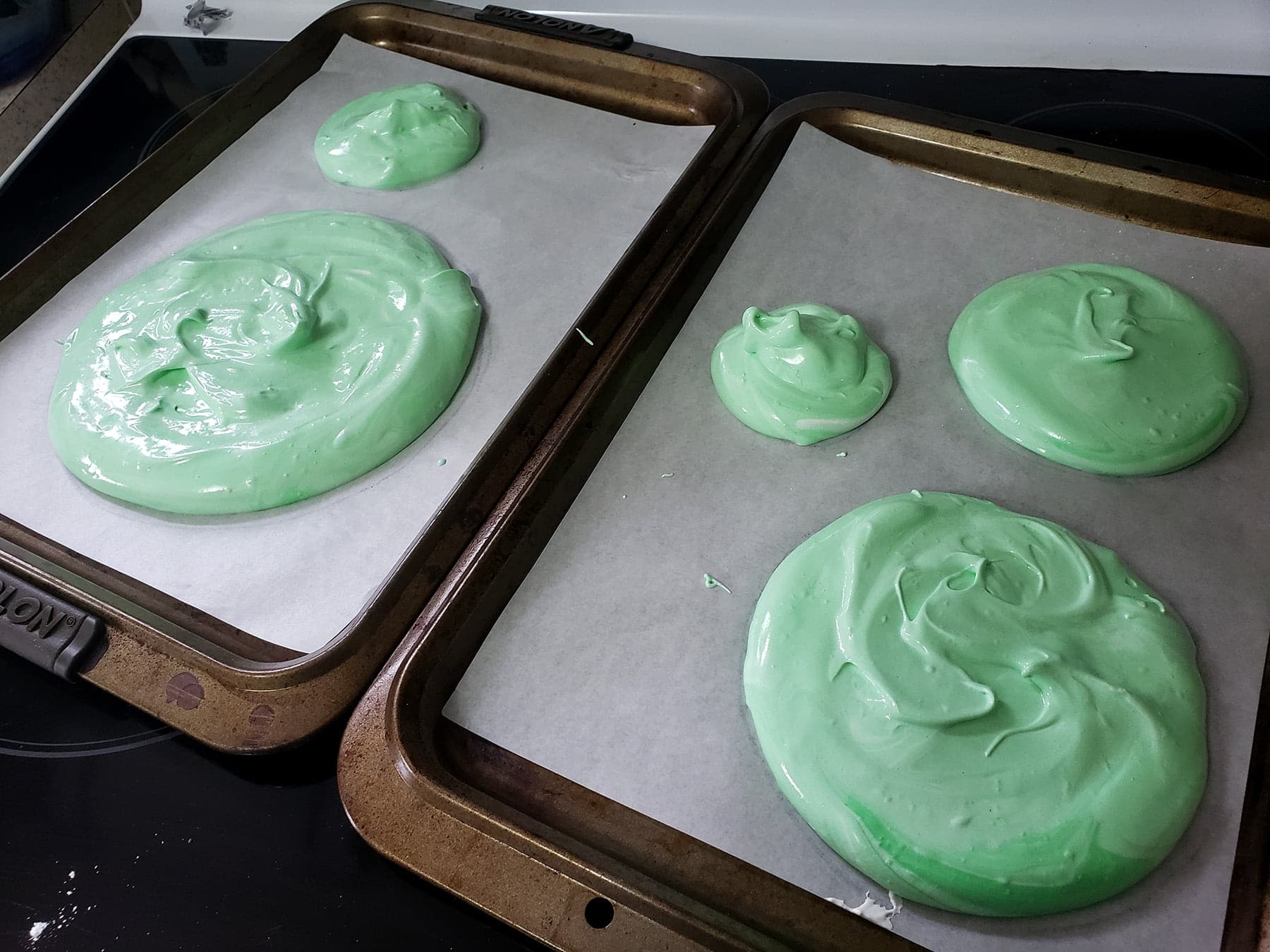 Two cookie sheets are lined with parchment paper, and have 5 rounds of green meringue - in various sizes - divided out between them.