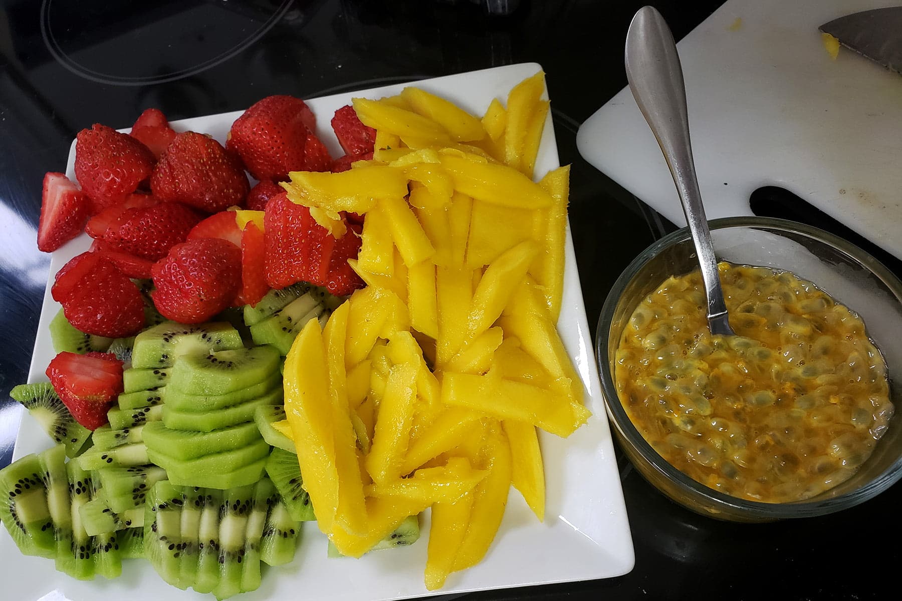 A plate of sliced mango, strawberry, and kiwi is resting next to a bowl of passionfruit guts.