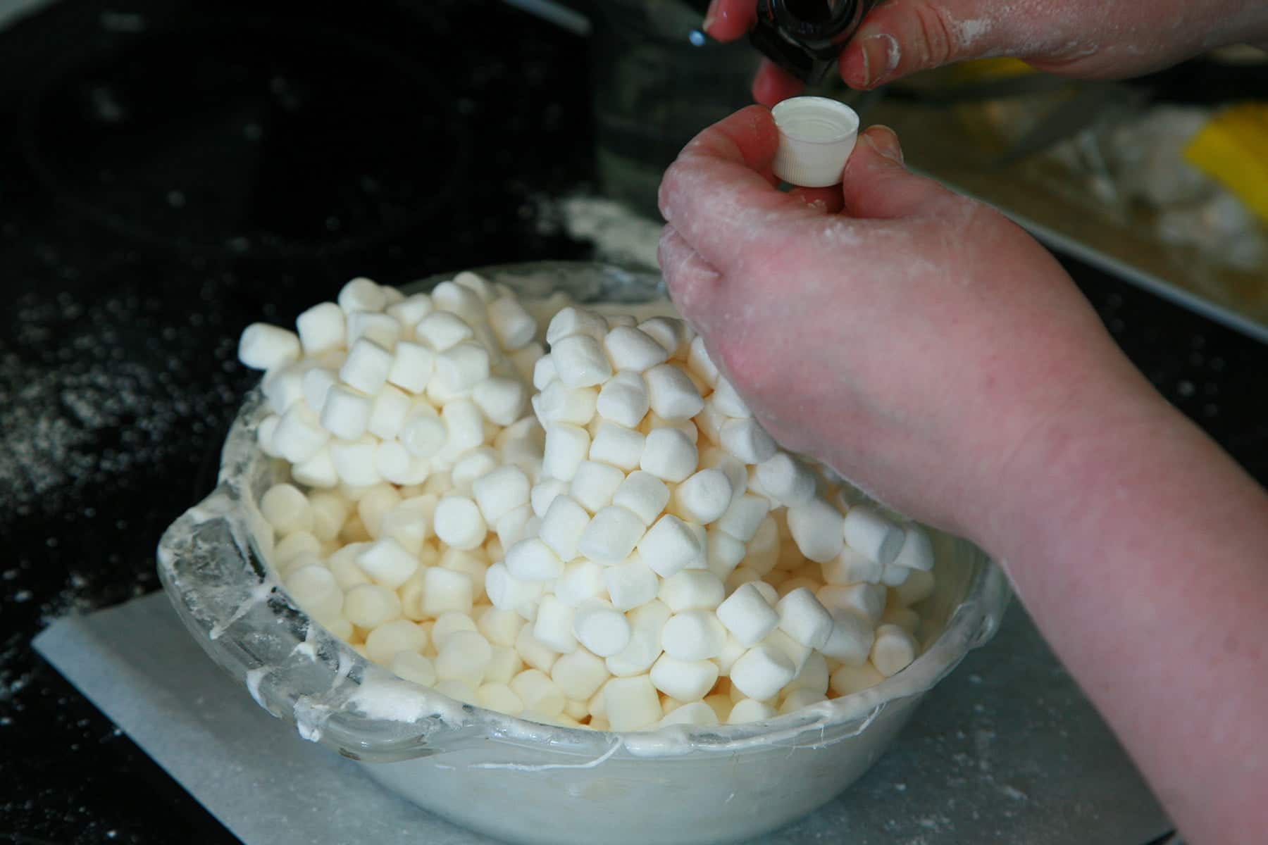 A glass bowl filled with mini marshmallows. A hand holds a cap of peppermint extract, about to add it to the mix.