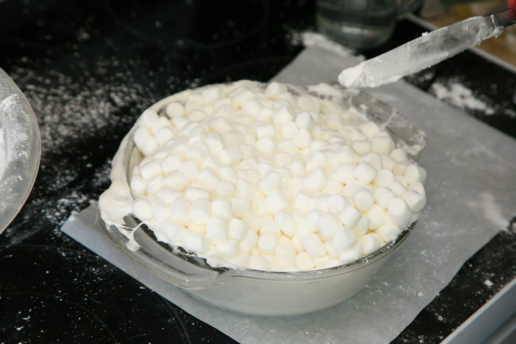 A glass bowl of melted mini marshmallows, being stirred with a knife.