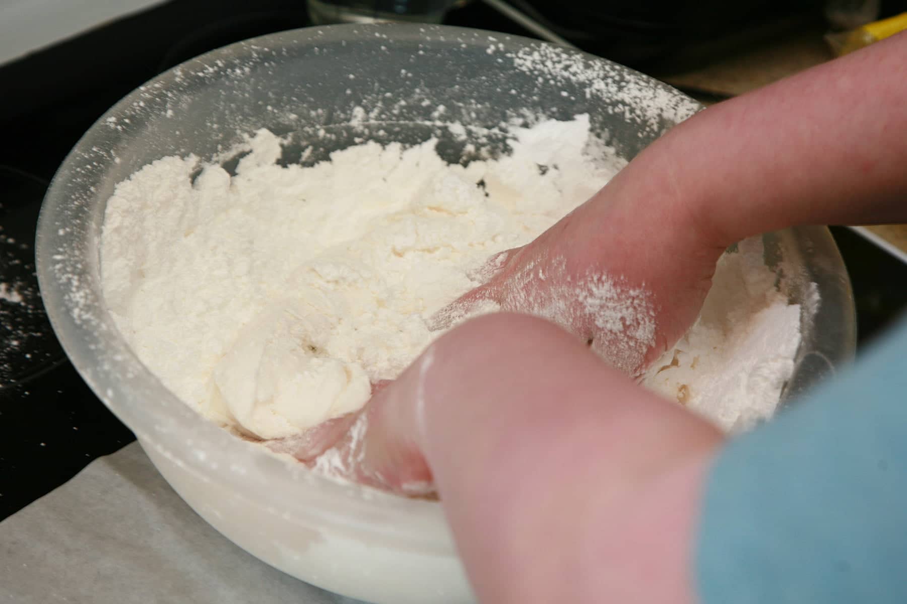 Two hands knead a mass of white marshmallow fondant in a large bowl of powdered sugar.