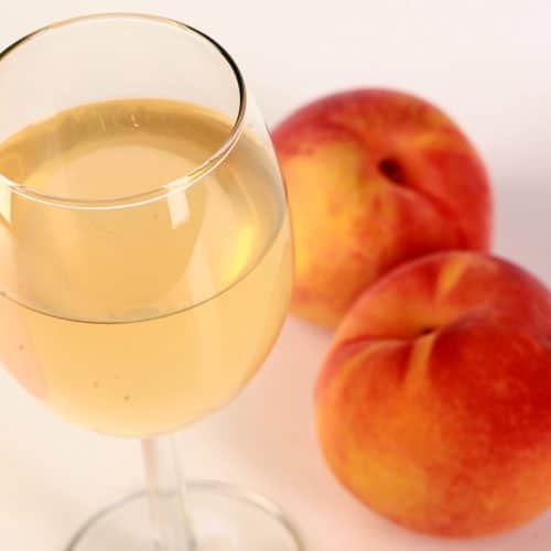 A glass of pale peach coloured wine, with 2 fresh peaches at the base of the glass.