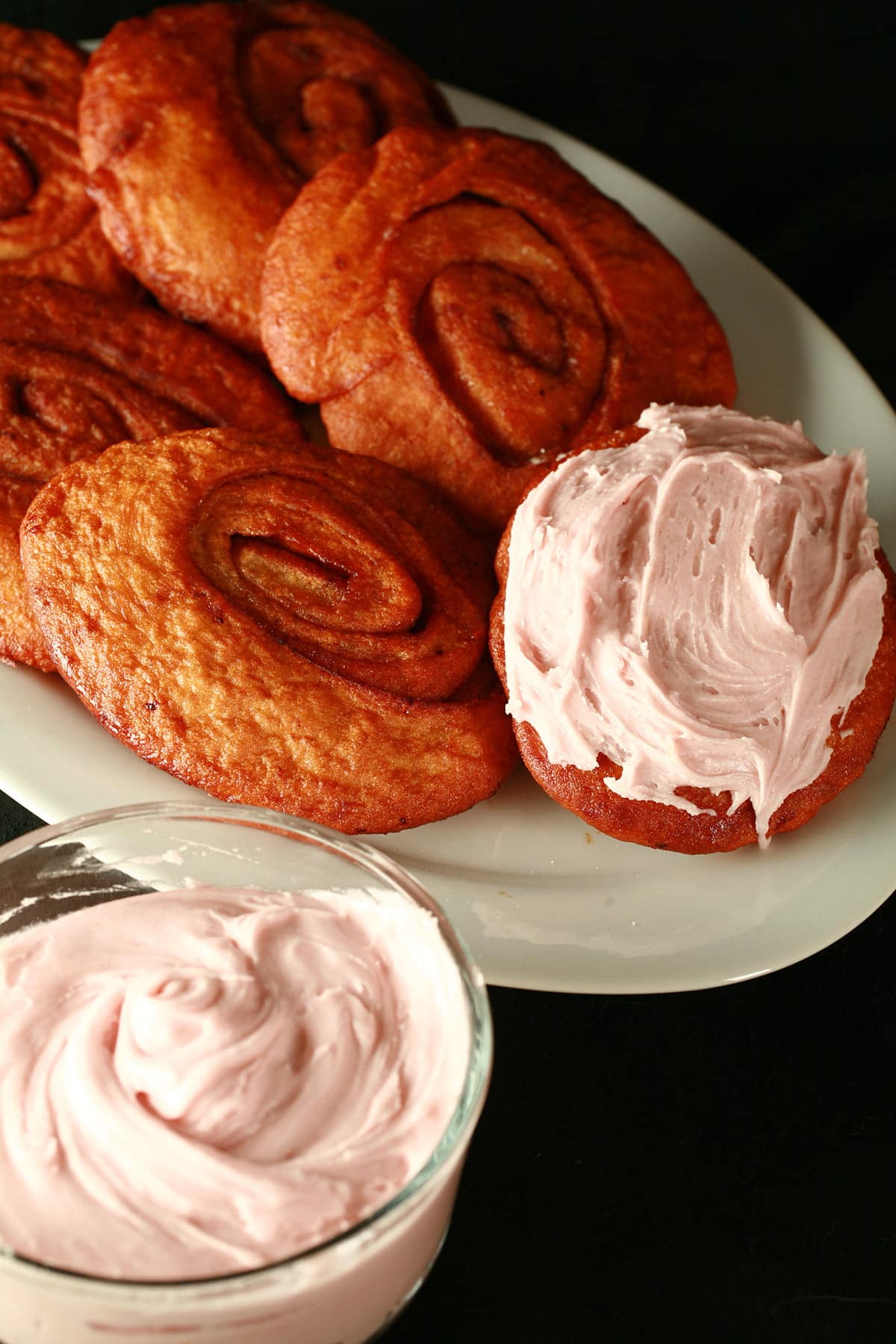Deep fried cinnamon rolls are arranged on a white platter, one is spread with a pink frosting - - made with this Persians recipe