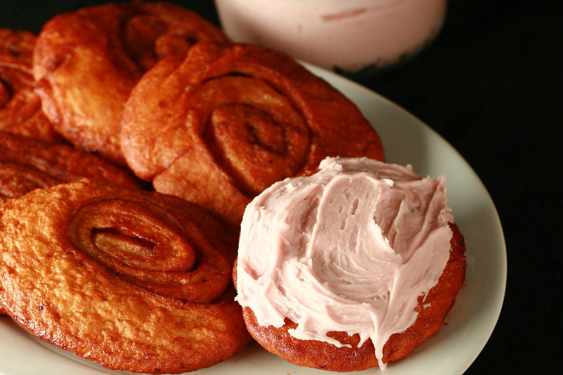 Deep fried cinnamon rolls are arranged on a white platter, one is spread with a pink frosting - A Persian roll!