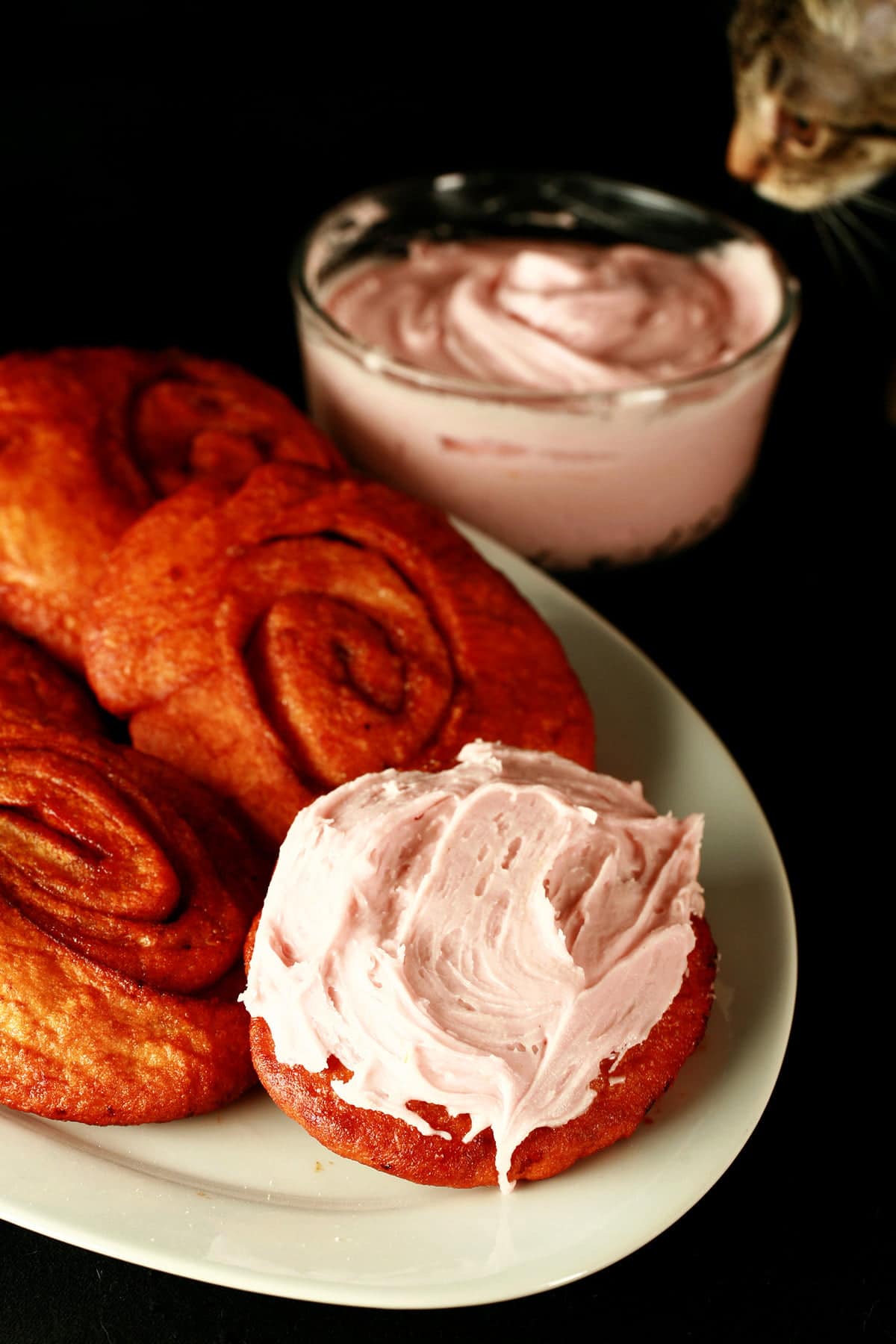 Deep fried cinnamon rolls are arranged on a white platter, one is spread with a pink frosting - A Persian roll! A grey tabby cat looks on.