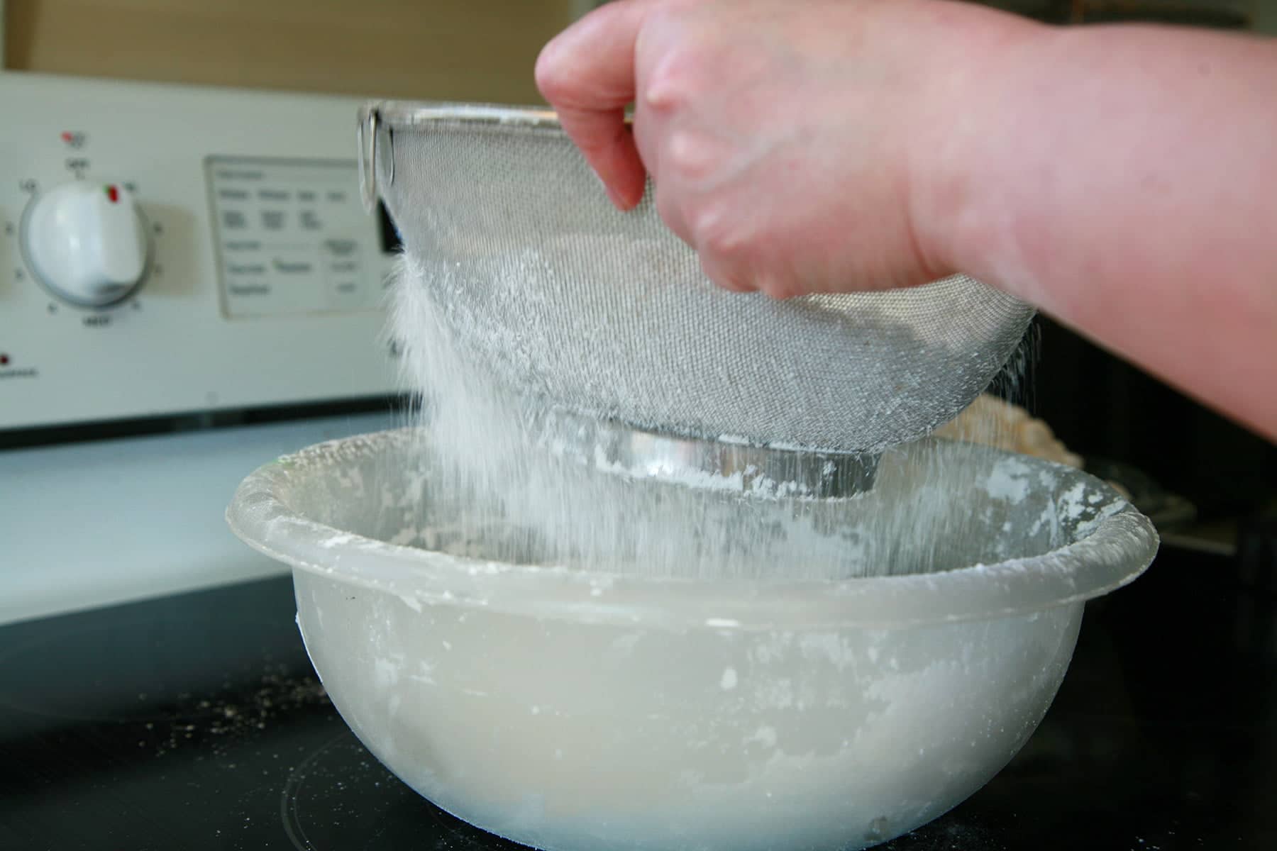 A hand holds a wire strainer over a large bowl, sifting powdered sugar into it.