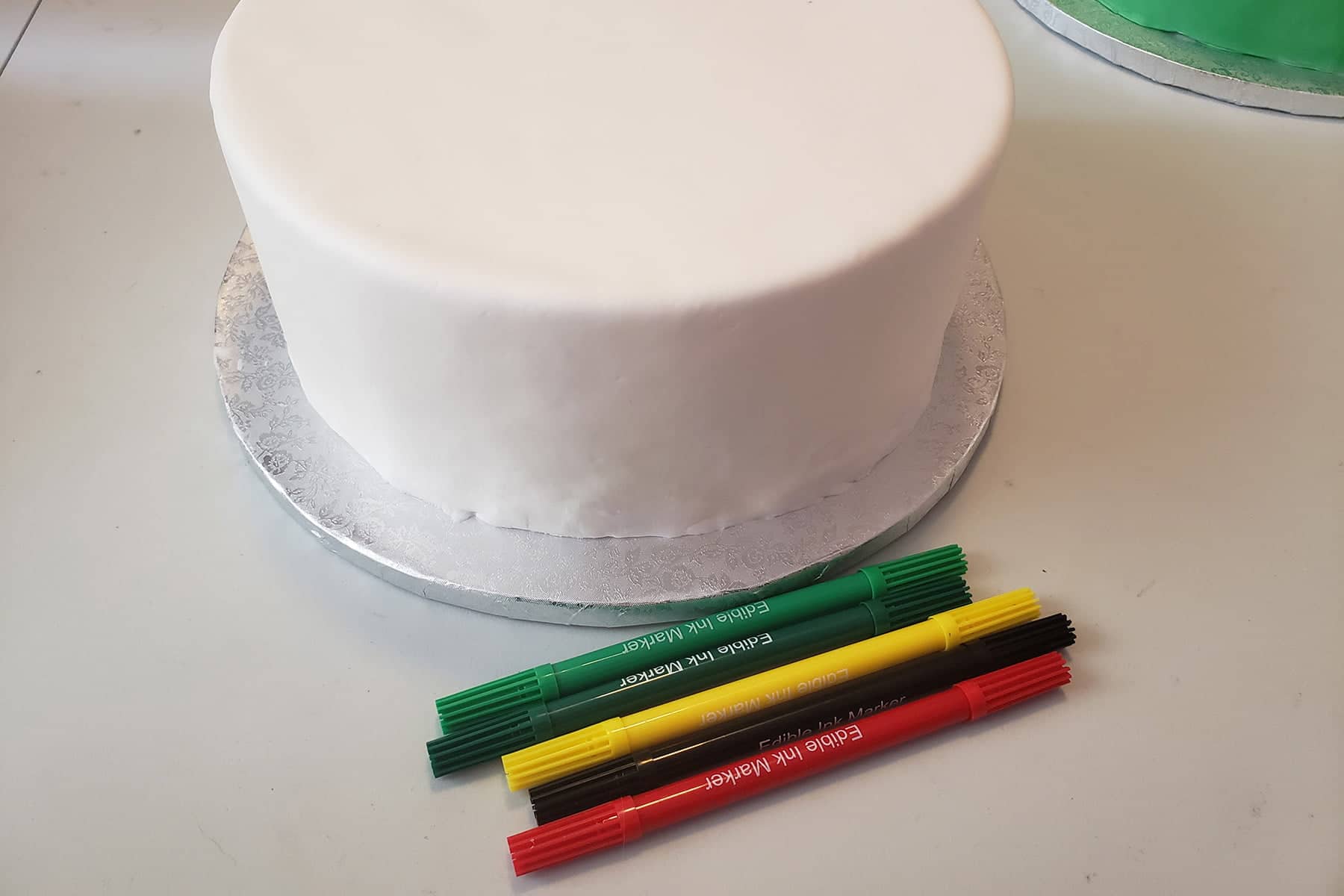 Bright green dark green, yellow, black and red food colouring markers are lined up in front of a large round cake covered in smooth white fondant.