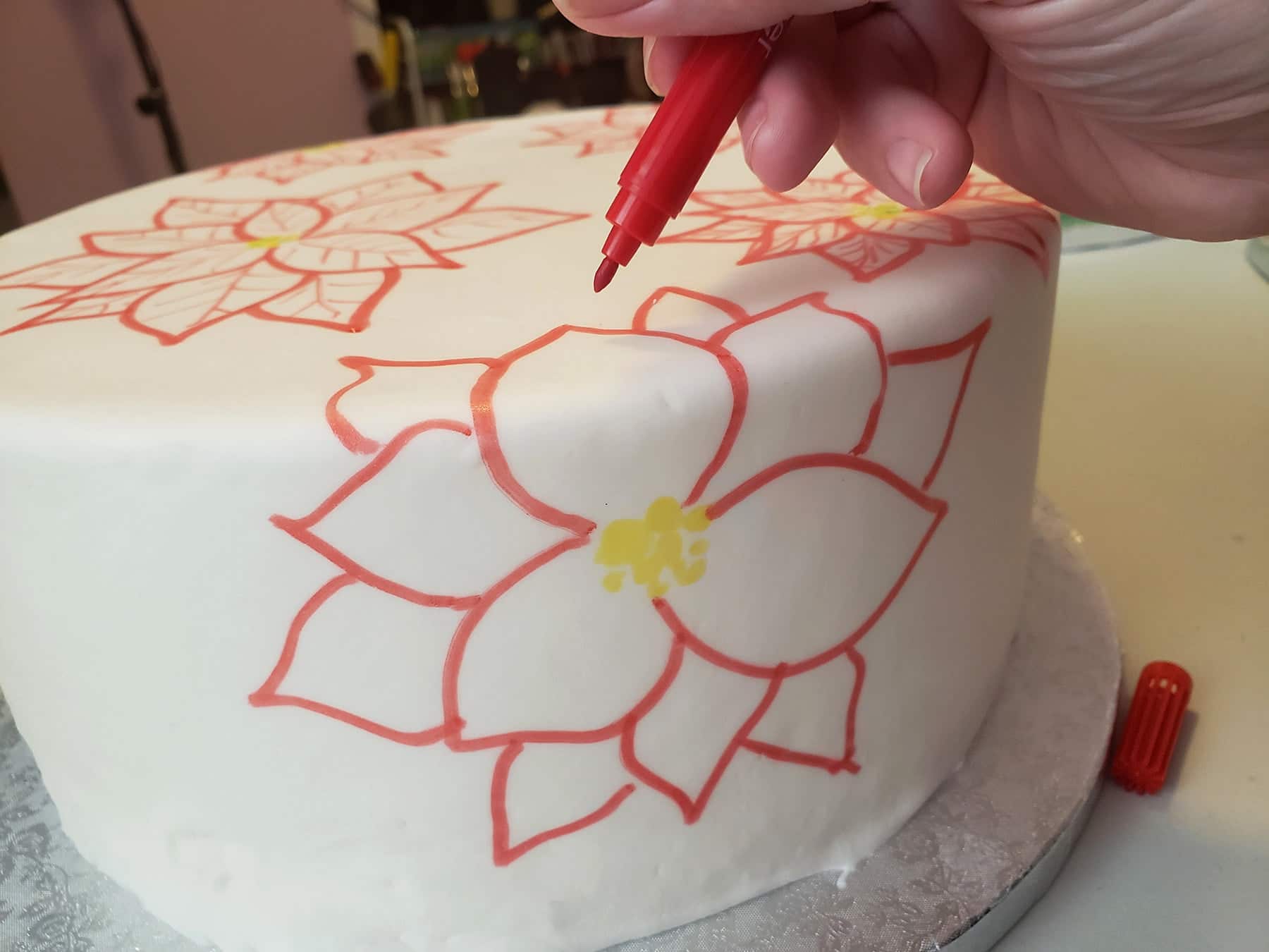 A hand uses a red food colouring marker to draw red poinsettias on a smooth white round cake.