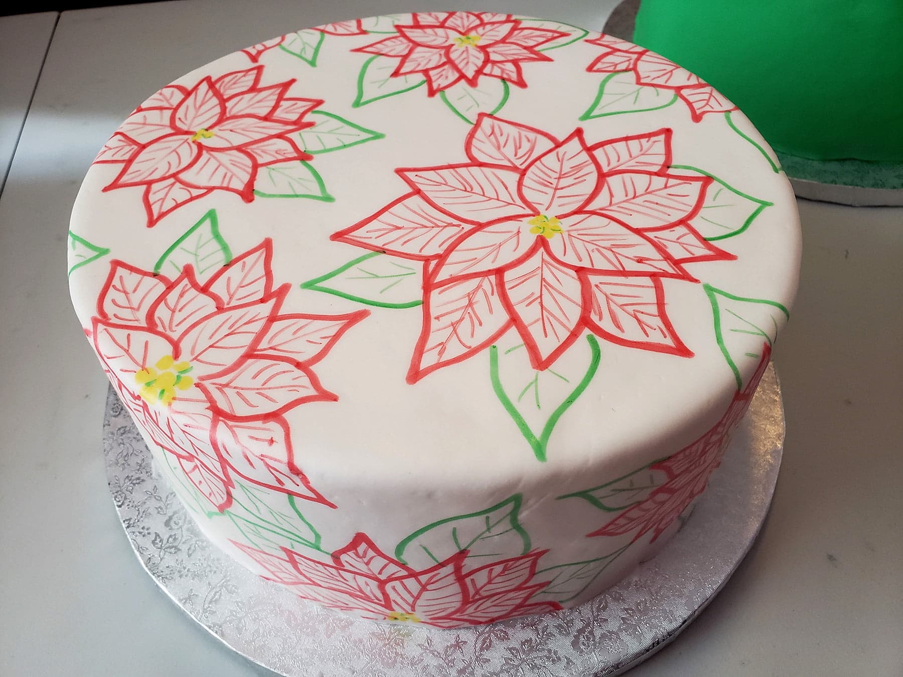 A large round cake covered in smooth white fondant has red poinsettias and green leaves drawn all over it.
