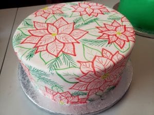 A large, round cake covered in smooth white fondant is covered in hand sketched poinsettias. The design is in red, 2 shades of green, and has yellow accents.