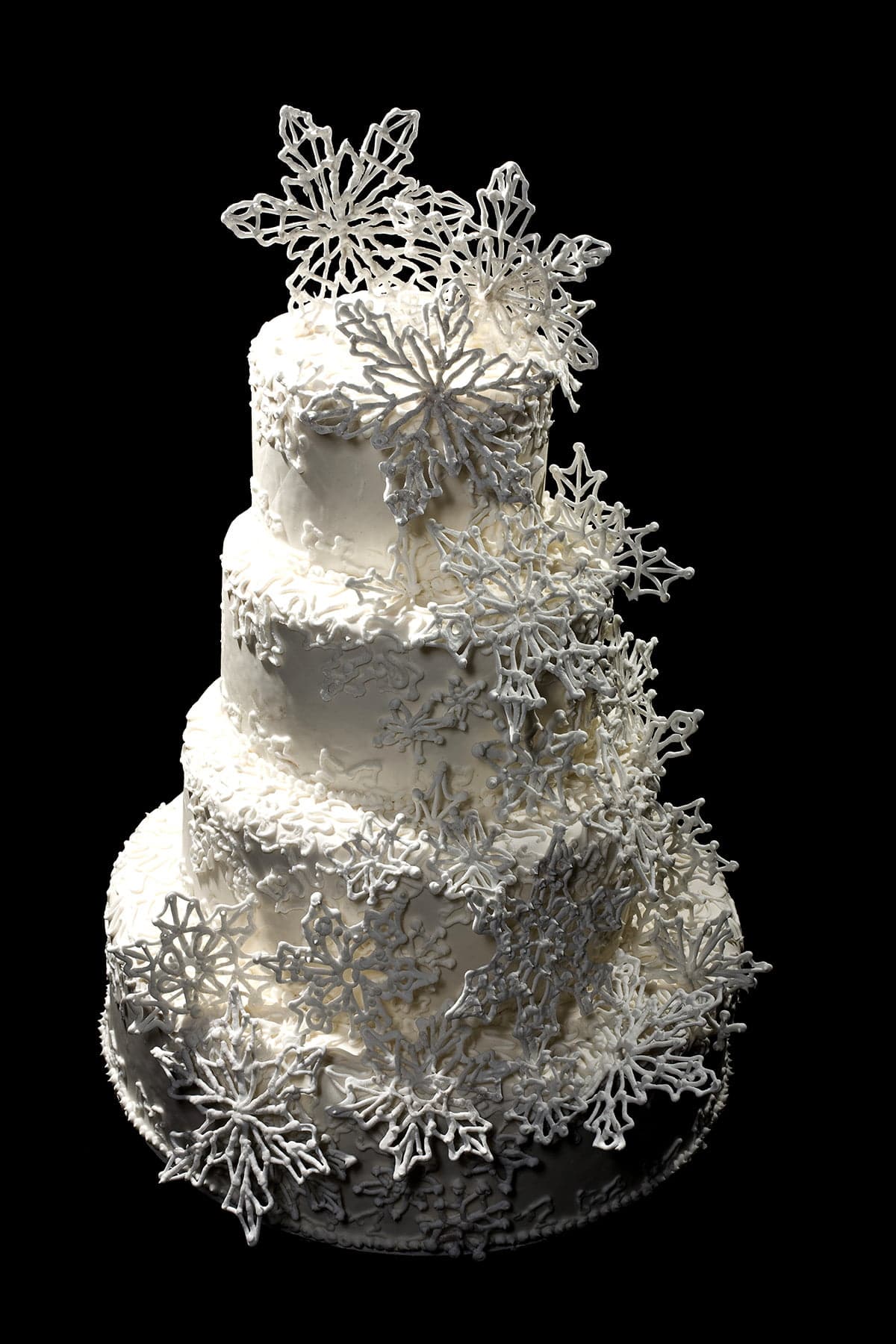Detail photo of a 4 tier white wedding cake, piped with an intricate lace design, and adorned with a cascade of 3D piped snowflakes.