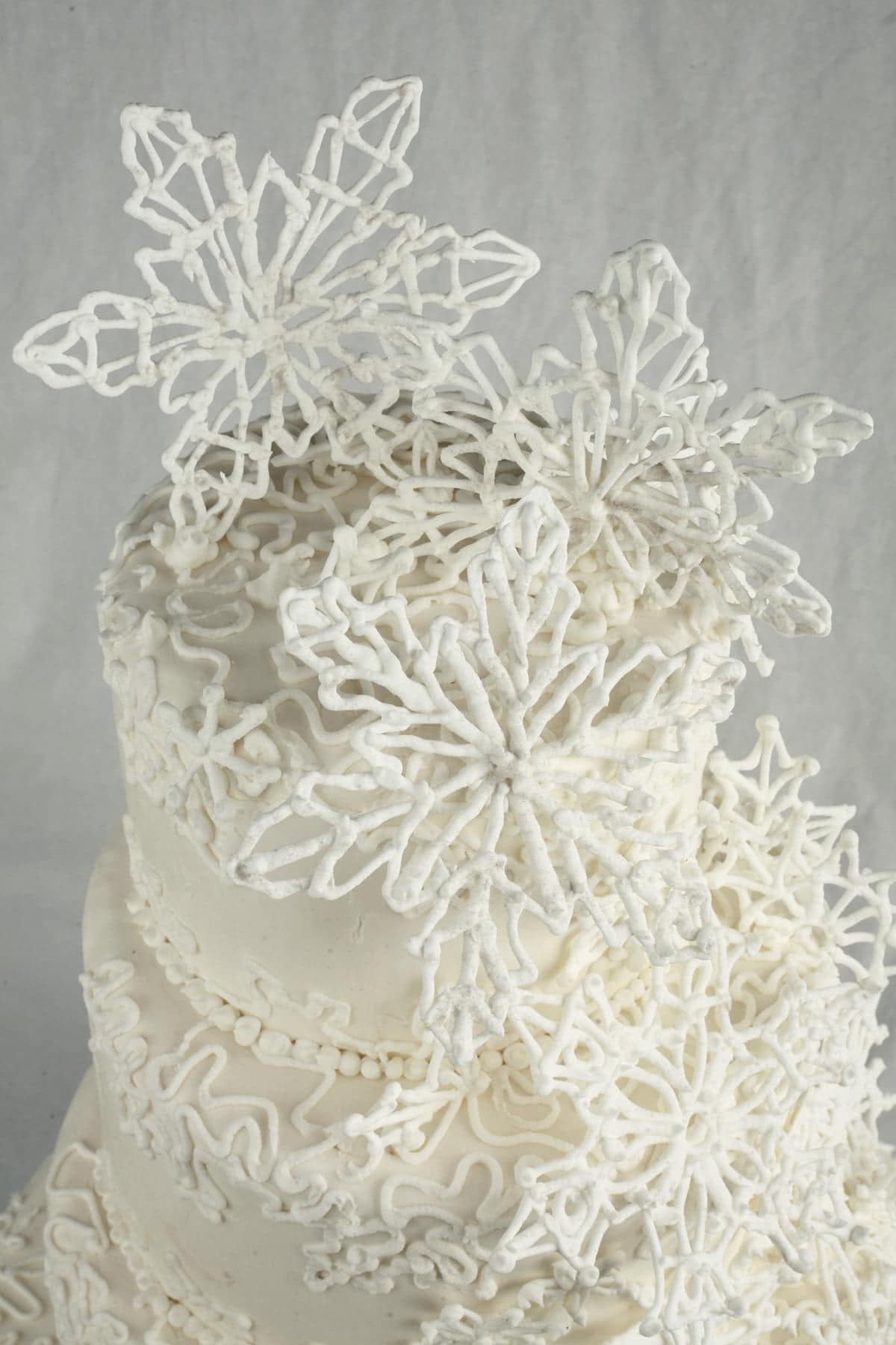 Detail photo of a 4 tier white wedding cake, piped with an intricate lace design, and adorned with a cascade of 3D piped snowflakes.