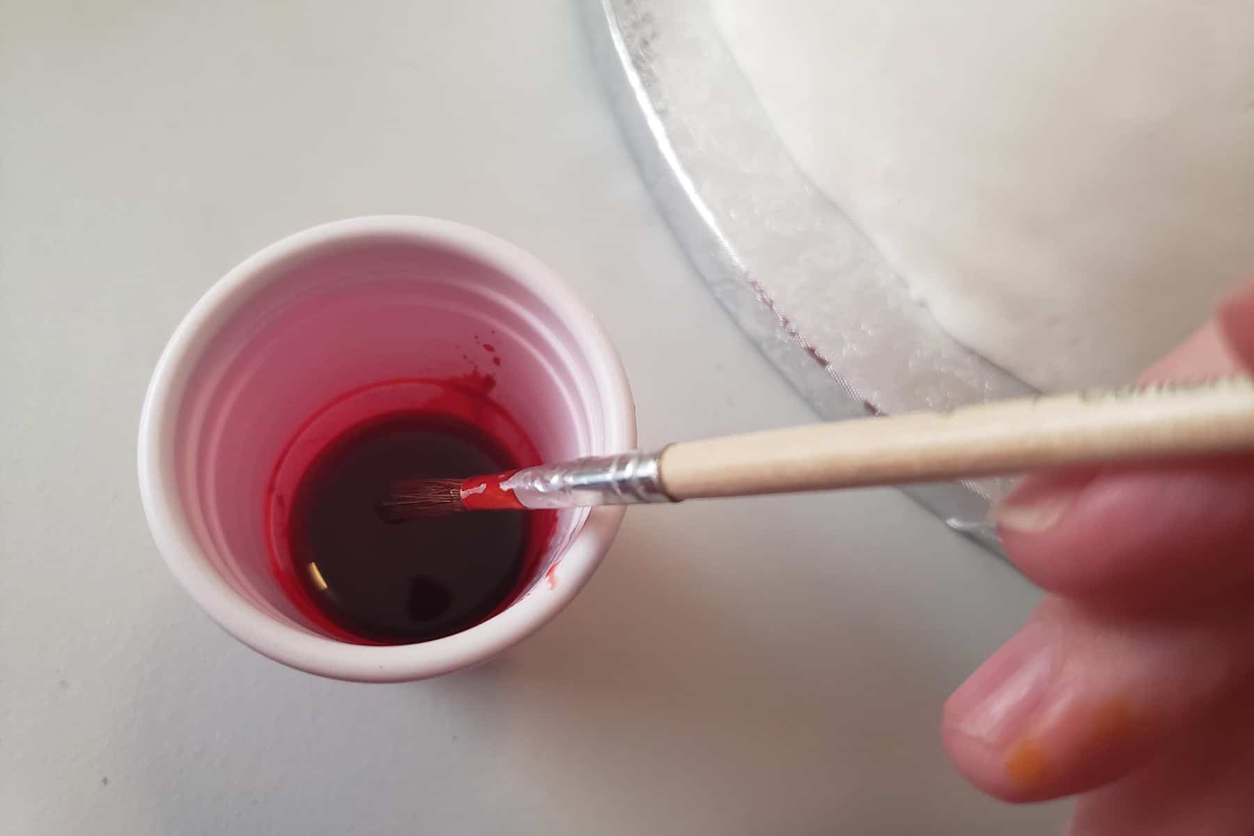 A small plastic cup with red foood colouring in it has a paintbrush being lifted out of it.
