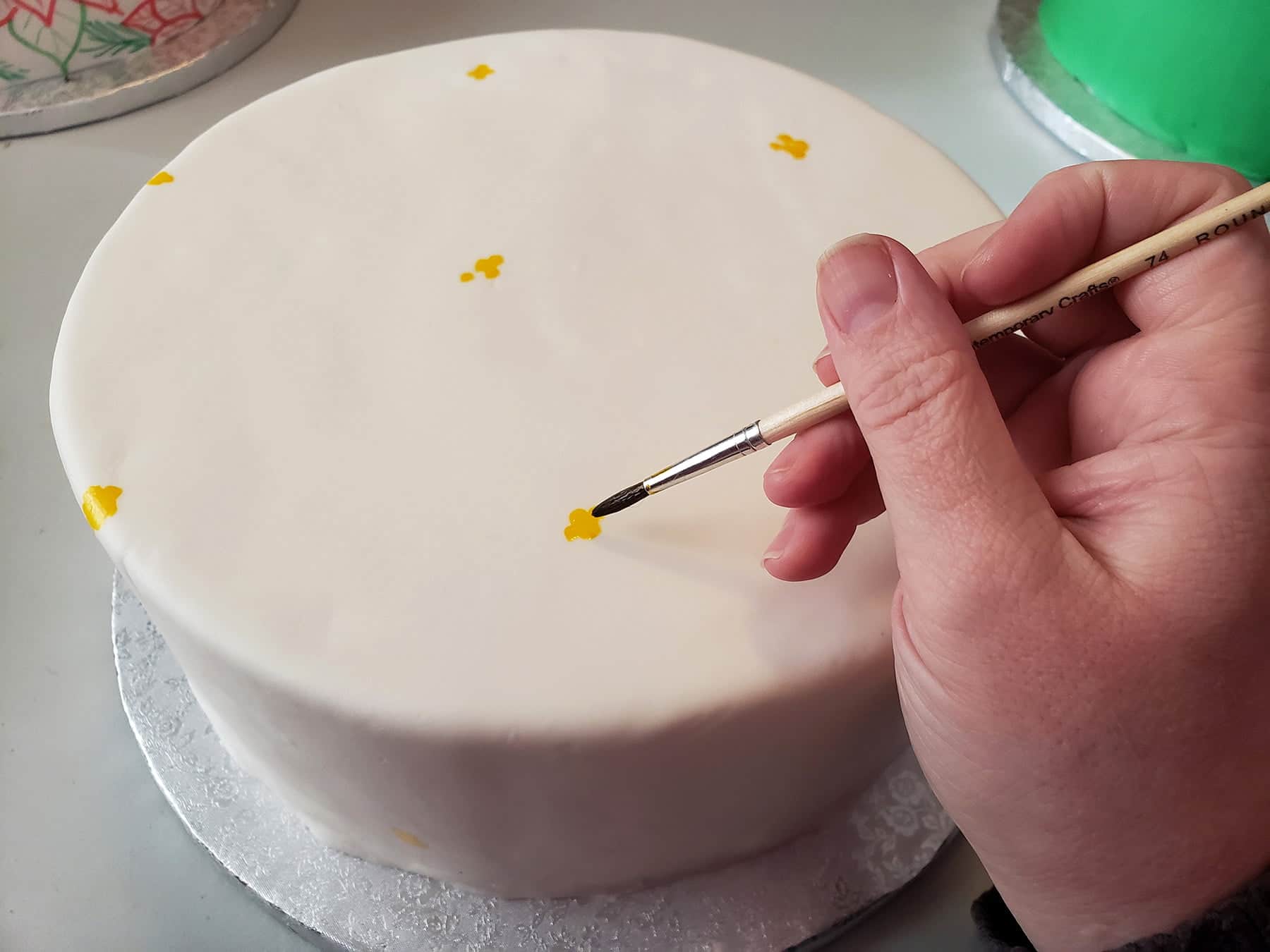 A hand uses a paintbrush to paint small dots of yellow food colouring on a smooth white round cake.