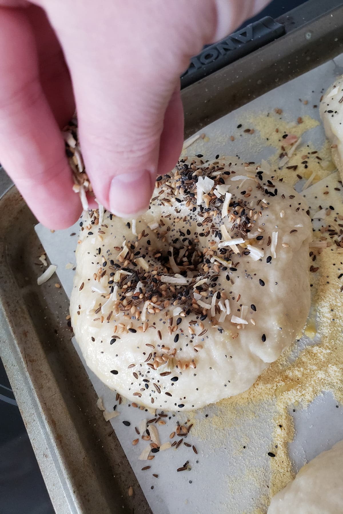 Fingers sprinkle "everything" seasoning over a raw bagel on a parchment lined baking sheet.