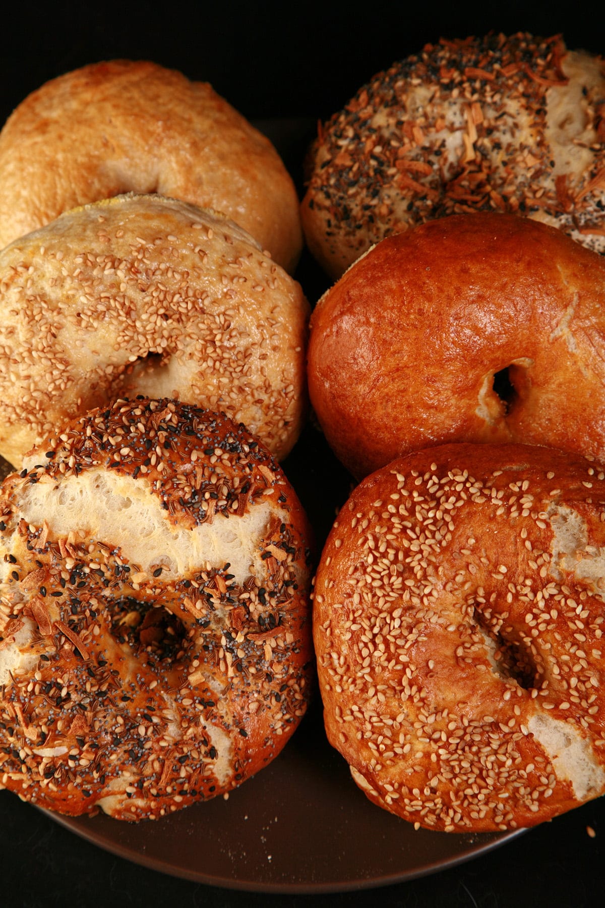Close up photo of a small brown plate, piled with 6 large homemade bagels, in various flavours.