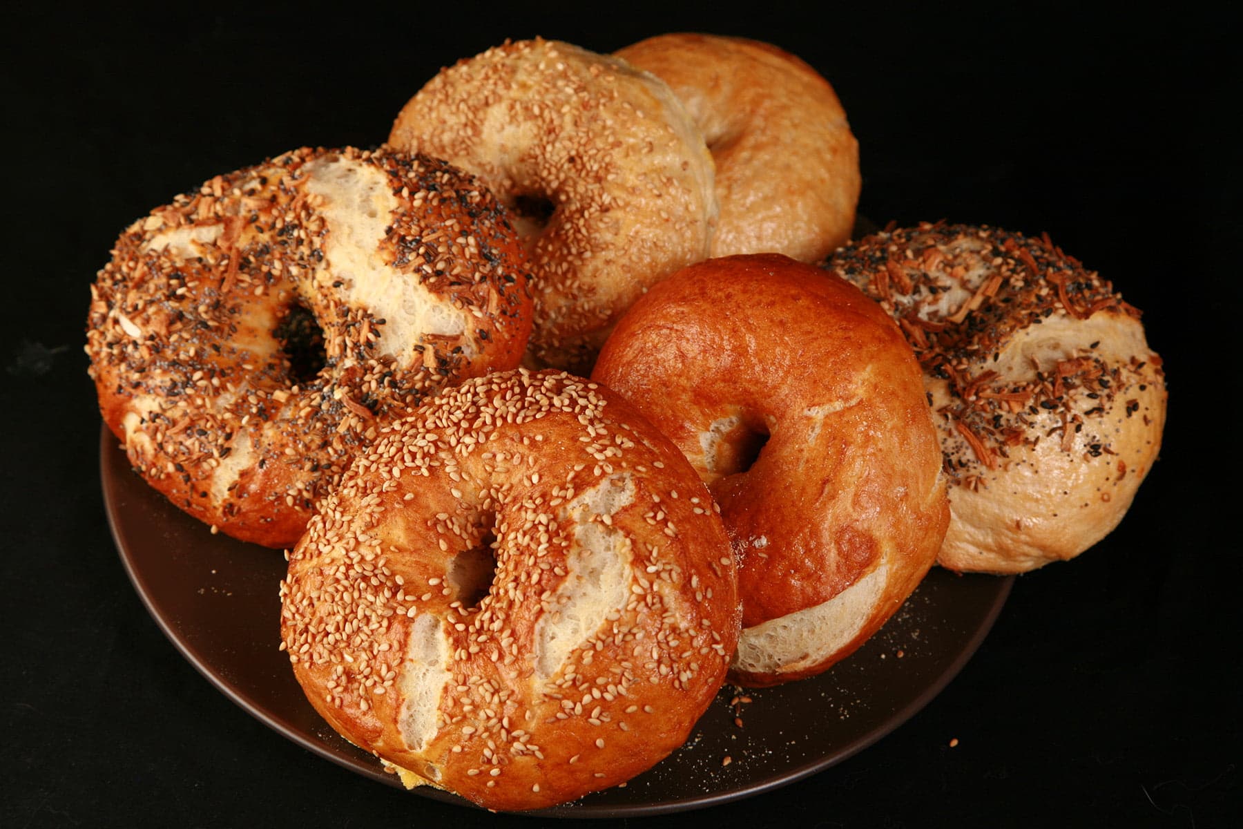 Close up photo of a small brown plate, piled with 6 large bagels, in various flavours. They were made with this How to Make Bagels tutorial.