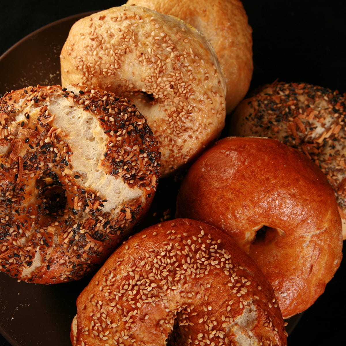 Close up photo of a small brown plate, piled with 6 large bagel, in various flavours. They were made with this bagel recipe.