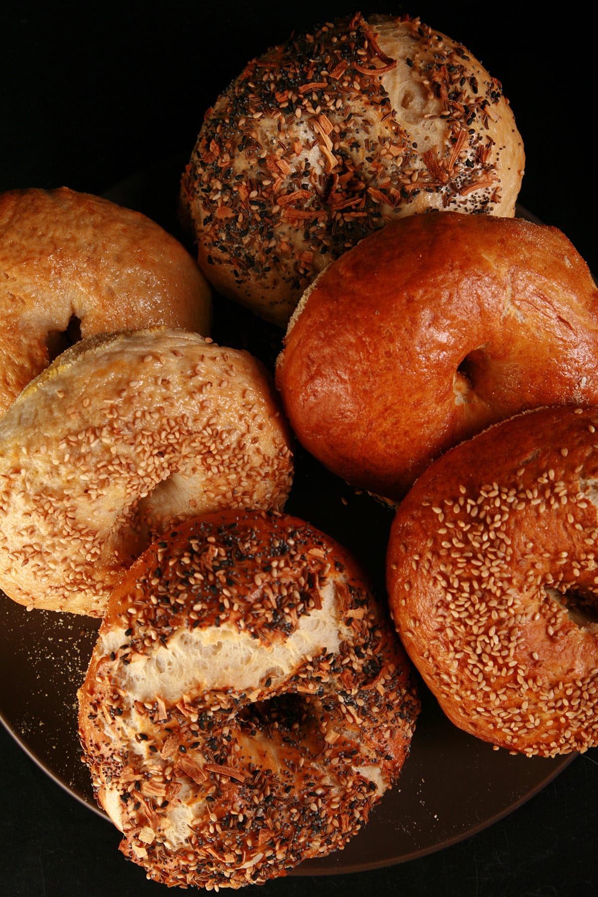 Close up photo of a small brown plate, piled with 6 large bagel, in various flavours. They were made with this bagel recipe.
