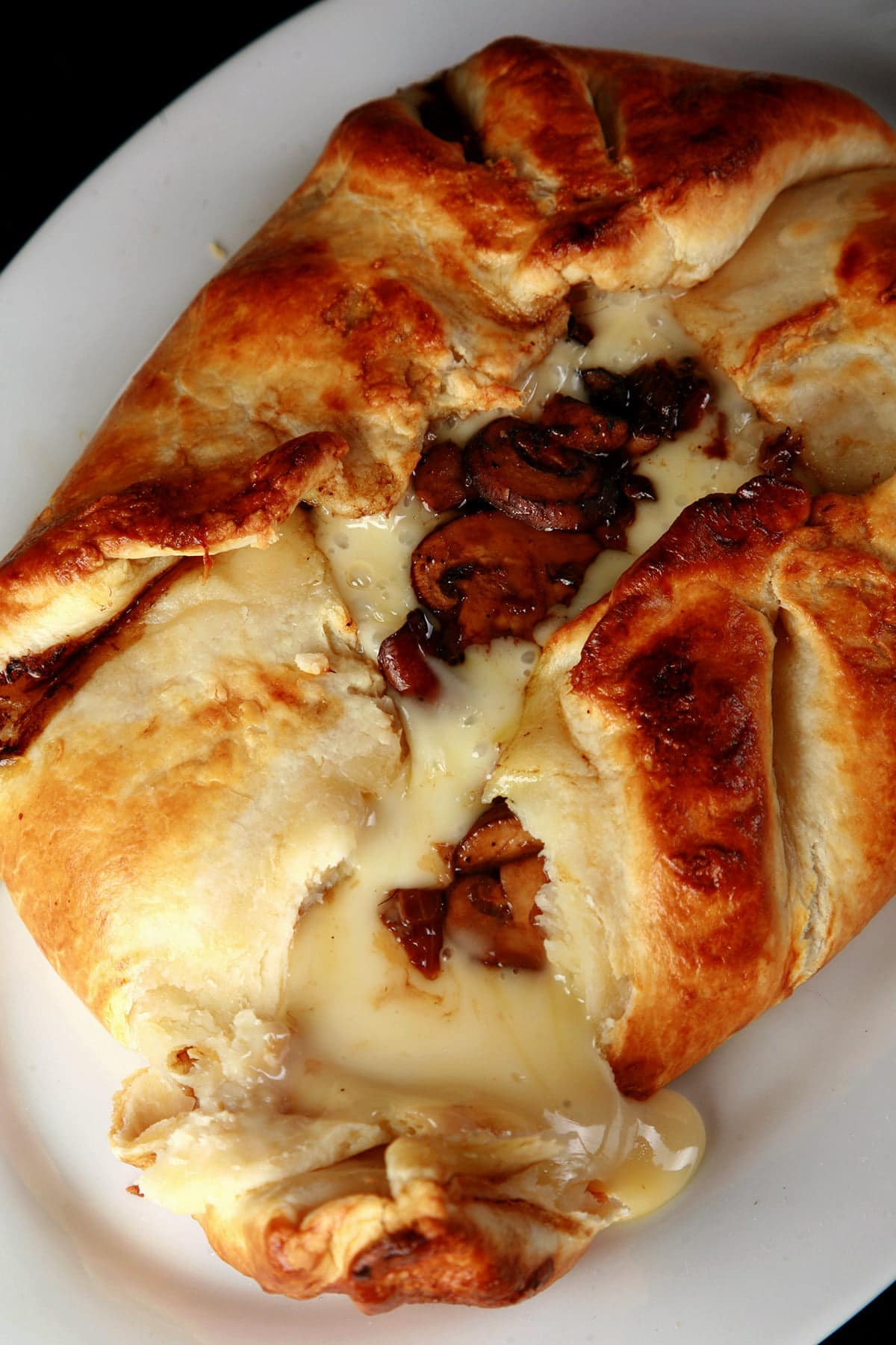 A balsamic mushroom baked brie is broken open, with melted cheese and sauteed mushrooms oozing out onto a white plate.