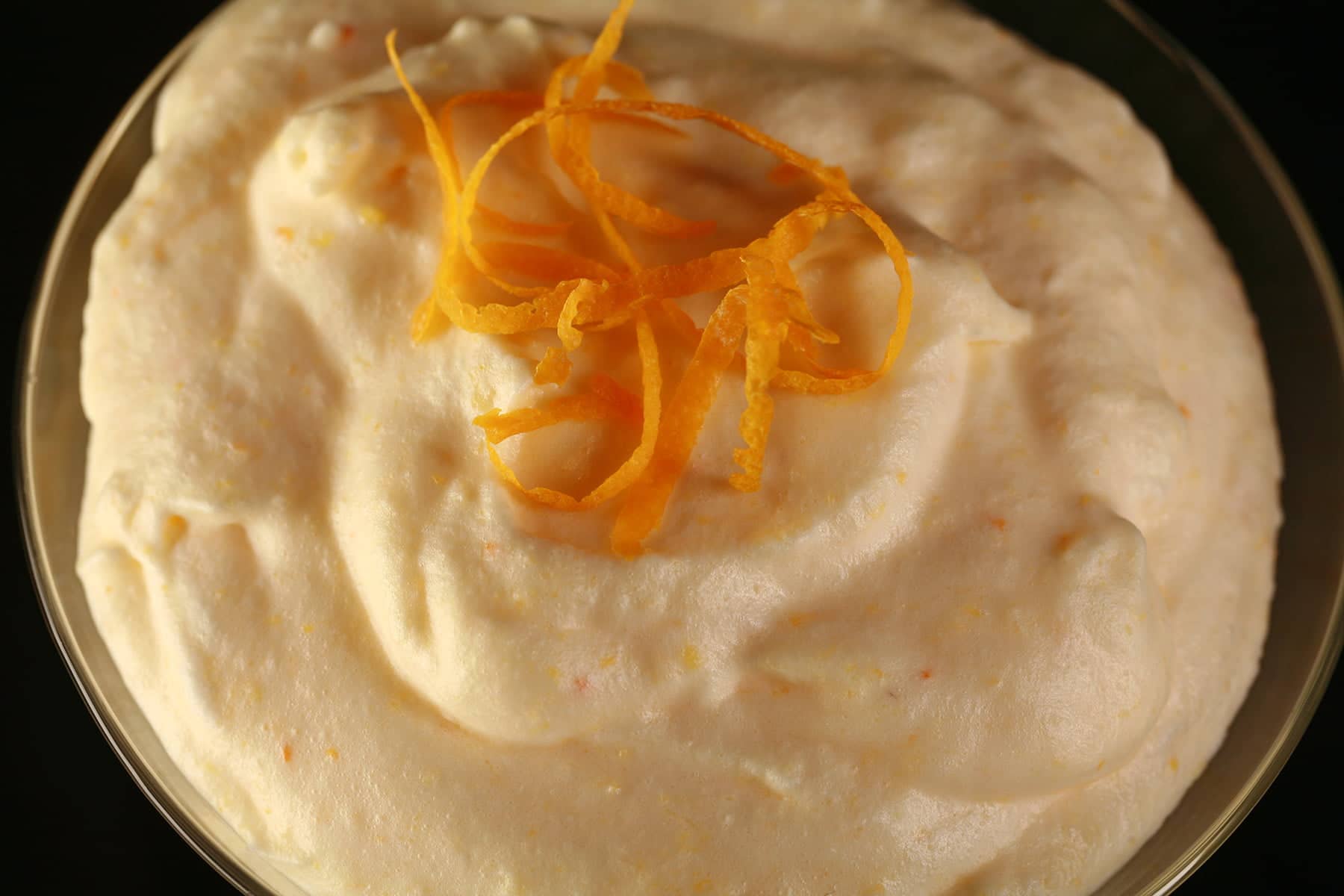 A very close up view of lightly orange coloured mousse. It has a small mound of clementine zest twists piled in the middle.