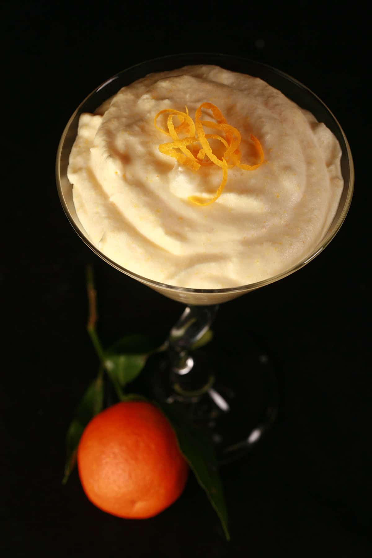 A martini glass is filled with an orange coloured clementine mousse. A clementine orange sits at the base of the glass.